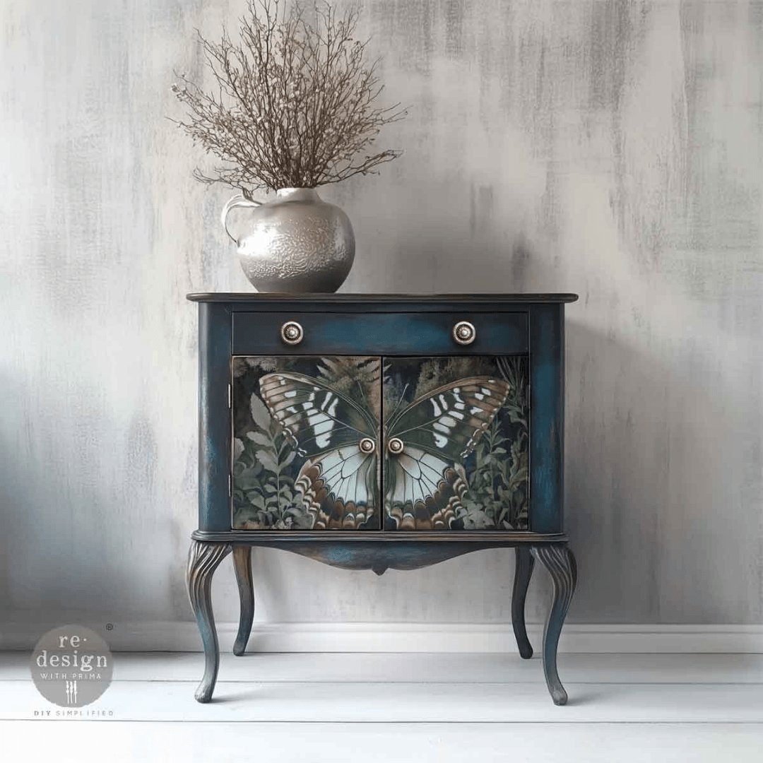 A small vintage-style side cabinet with a dark blue painted frame and butterfly artwork on the doors, styled with a silver vase of dried branches on top against a textured gray wall.