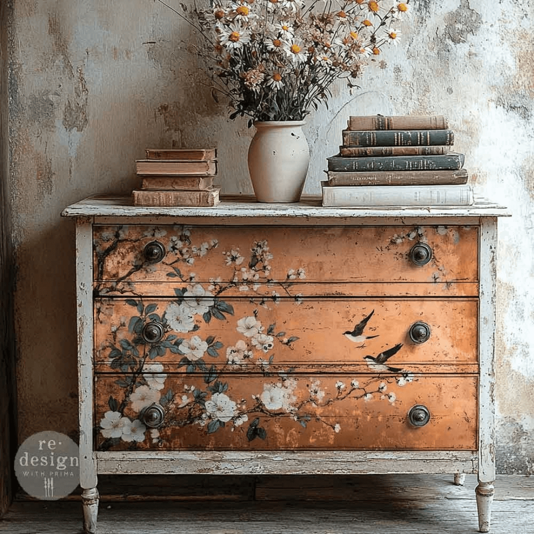 Vintage dresser with floral and bird design, books, and a vase on top against a textured wall.