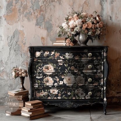A vintage-style black wooden chest of drawers with floral decals, styled in a rustic room with a distressed plaster wall, surrounded by books and bouquets of pink and peach roses.
