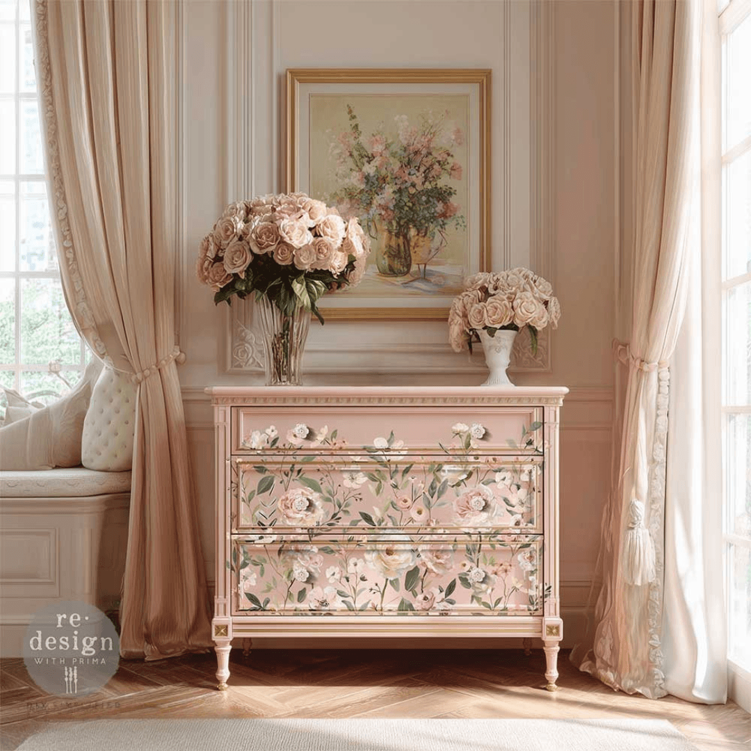 A pale blush pink three-drawer dresser with floral painted detailing, styled in a classic French-chic room with beige curtains, a window seat, and bouquets of pink roses.