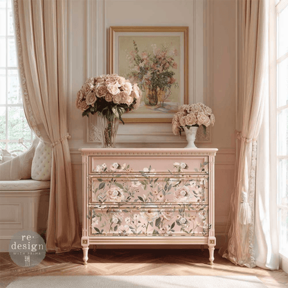 A pale blush pink three-drawer dresser with floral painted detailing, styled in a classic French-chic room with beige curtains, a window seat, and bouquets of pink roses.