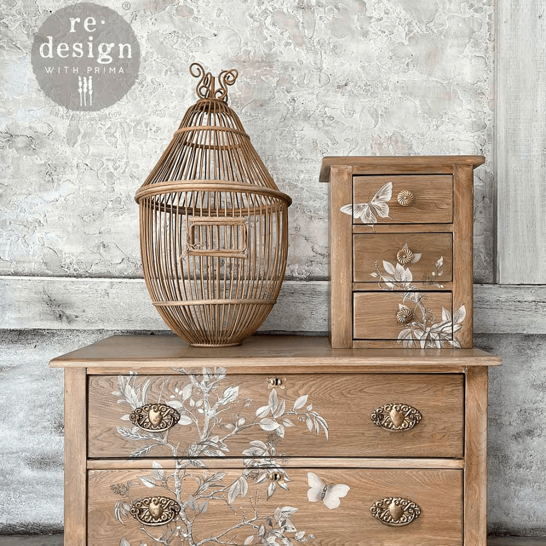 A styled interior scene showing a wooden chest of drawers with floral decals, a matching small three-drawer cabinet, and a decorative rattan birdcage. 