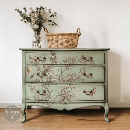 A vintage-style three-drawer chest of drawers painted in a soft sage green with delicate bird and floral illustrations across the front. It is styled in a room setting with a wicker basket and a vase of dried flowers on top, set against a plain off-white wall and light wood floor.
