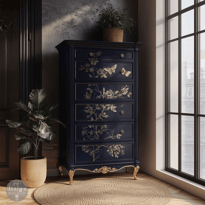 A tall dark navy blue five-drawer chest of drawers with ornate gold floral and butterfly motifs on the drawer fronts, styled in a room next to a window with potted plants.