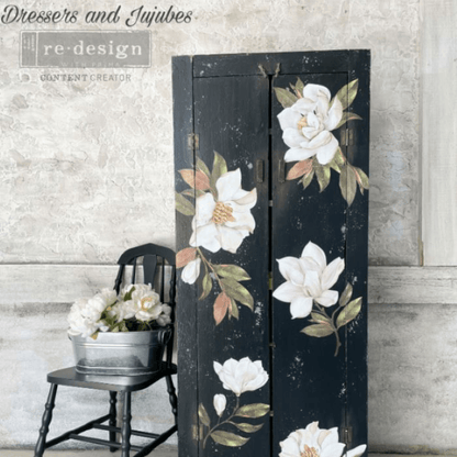 A tall black wooden cabinet or screen decorated with large white magnolia flowers and green leaves, styled indoors against a textured light gray wall next to a small black chair with a bucket of flowers. 