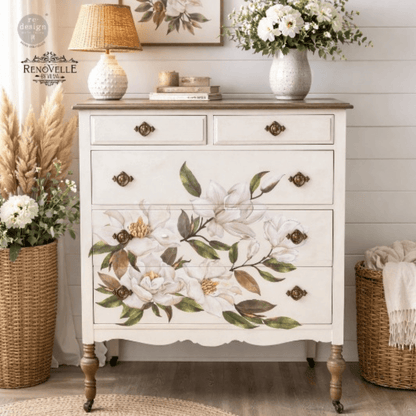 A white three-drawer dresser with a painted magnolia floral design on the front, styled in a bright, rustic room with a lamp, framed art, and wicker baskets.