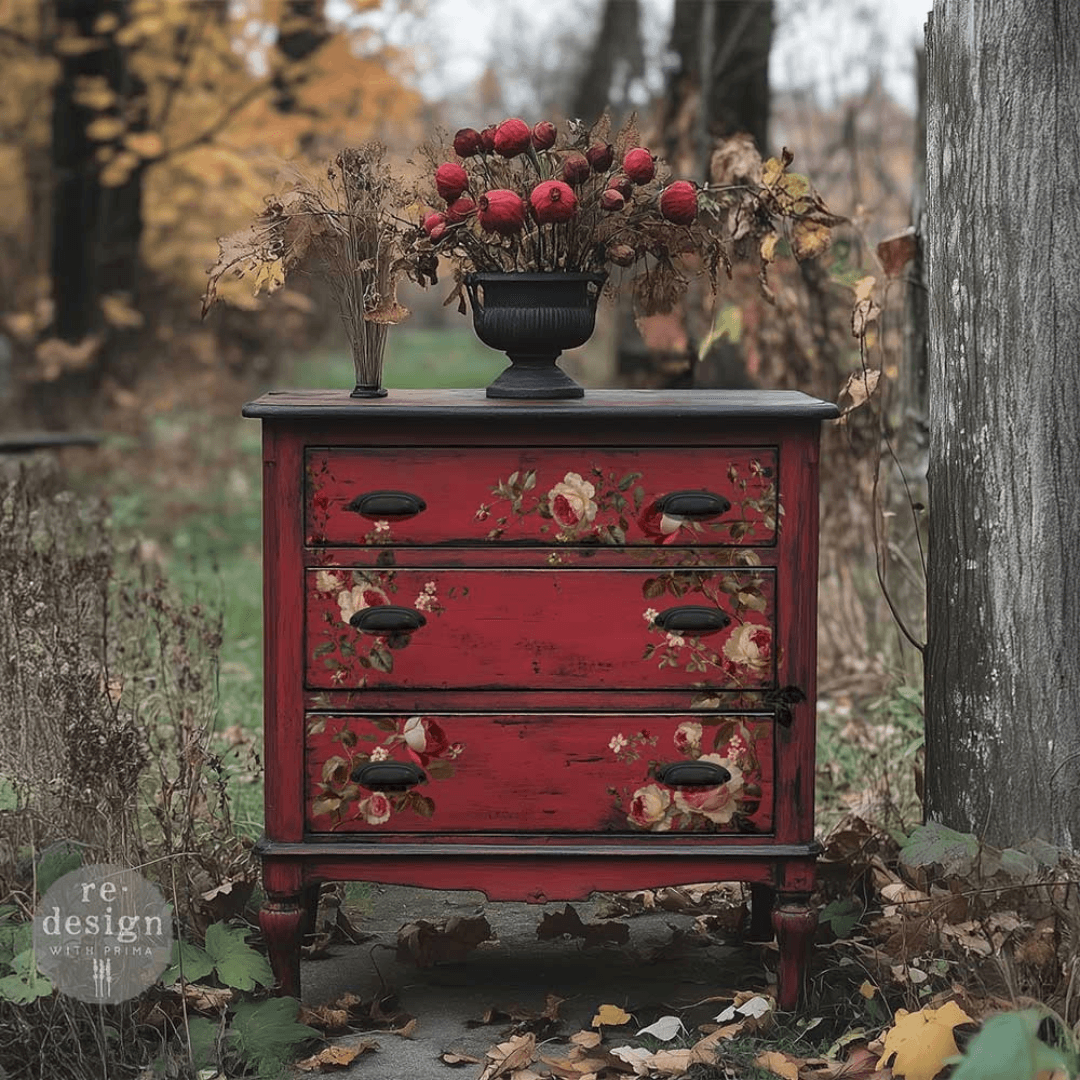 A distressed red three-drawer dresser with floral decals and dark metal handles, styled outdoors on a stone path with autumn leaves, a tree trunk, and dried floral arrangements on top.